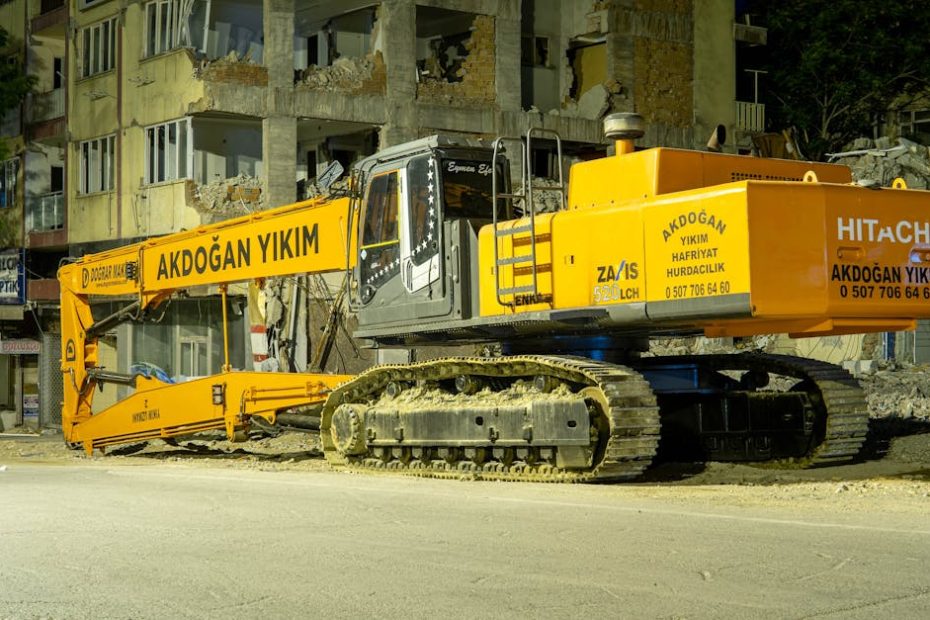 Large excavator at a city demolition site during nighttime, illuminated by streetlights.