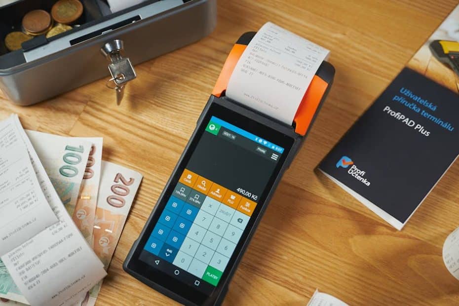 A modern payment terminal with receipts, cash, and manual on a wooden table.