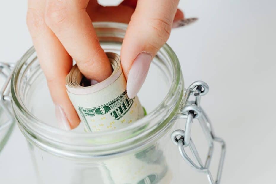 A close-up of a hand placing rolled dollars into a glass jar, symbolizing savings.