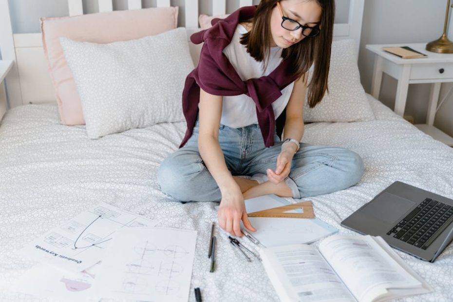 A young woman studying on her bed with a laptop, books, and papers scattered around.