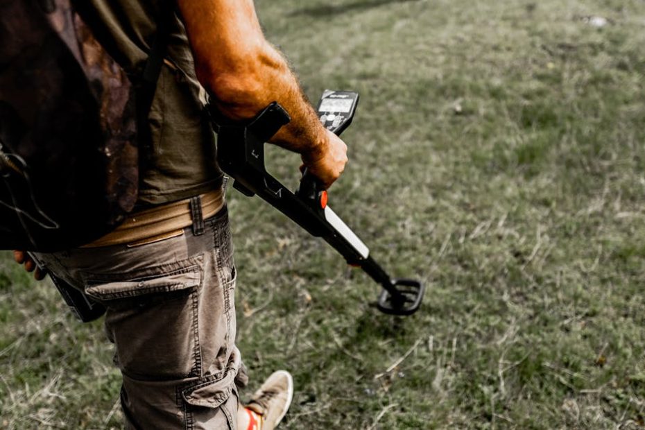 Person using a metal detector on a grassy field wearing casual attire.