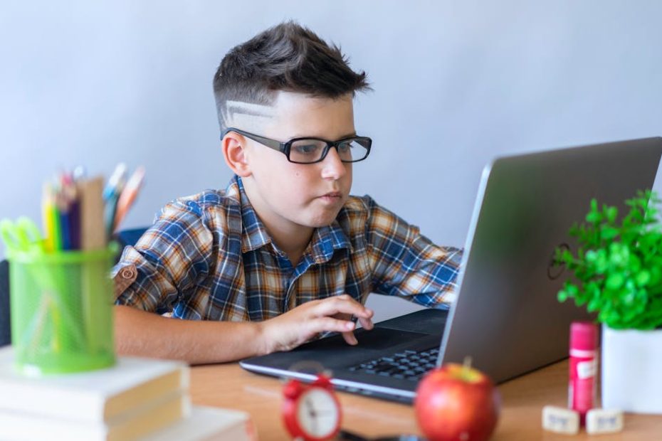 A young boy wearing glasses focused on using a laptop for online learning, surrounded by school supplies.