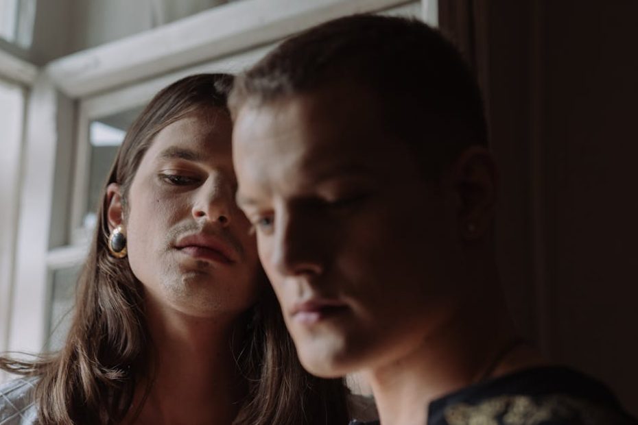 Dramatic indoor portrait of two androgynous individuals by a window, evoking mystery and sophistication.