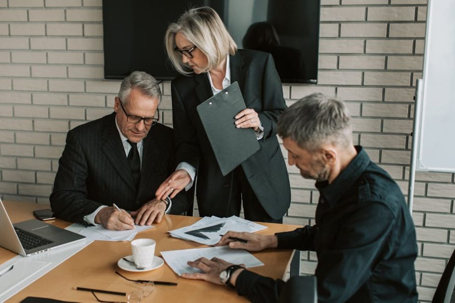 Three mature professionals in a business meeting discussing and signing documents in an office setting.