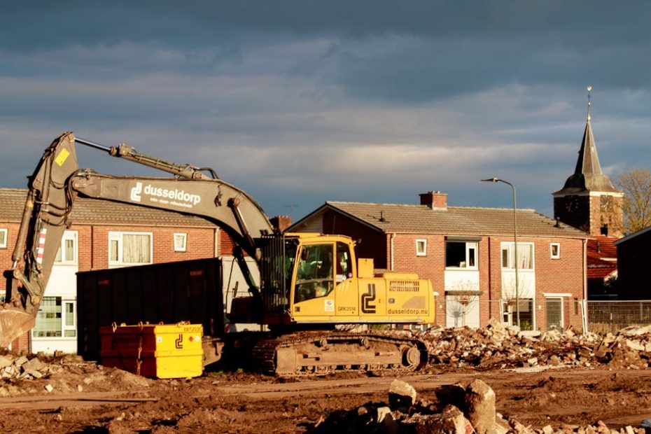 Excavator at a construction site near residential houses under a dramatic sky.