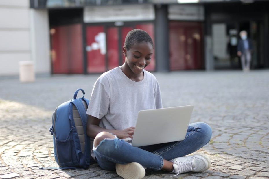 Smiling woman sitting on cobblestone street with laptop, engaging in online learning outdoors.